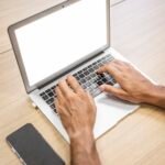 digital: Un gros plan des mains d'une personne tapant sur un ordinateur portable avec un smartphone à proximité sur un bureau en bois, symbolisant la révolution numérique de 2026 en Côte d'Ivoire qui comble le fossé de communication entre le gouvernement et ses citoyens.